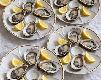 Op de afbeelding: Drie witte keramische oesterborden met goudkleurige randen, elk voorzien van voorgevormde uitsparingen voor zes oesters. Elk bord is opgemaakt met verse rauwe oesters en heldergele partjes citroen, plus twee extra citroenhelften.