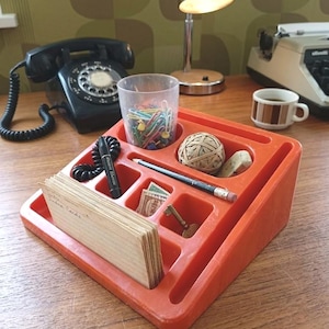 May include: A bright orange plastic desk organizer with multiple compartments containing a stack of index cards, a clear cup of colorful paper clips, a rubber band ball, a pencil, a black pen, a small brass key, postage stamps, and an eraser.