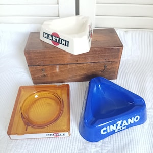 May include: A collection of three ashtrays including a triangular white ceramic ashtray with the Martini logo, a square amber glass ashtray with a Martini label, and a triangular blue ceramic ashtray with the Cinzano Blanc logo.