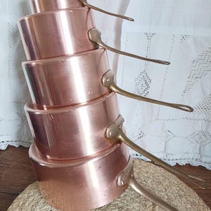 Può includere: Una pila di cinque pentole di rame con manici in ottone. Le pentole sono di diverse dimensioni e hanno una superficie lucida e riflettente. I manici sono lunghi e curvi, fissati con rivetti. Sono disposte su un tappetino intrecciato.