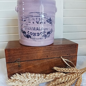 Op de afbeelding: Een lavendelkleurige keramische pot met twee kleine handvatten en een verweerd zwart etiket met de tekst "Finest Orange Marmalade London". De pot staat op een houten kist. Een bosje tarwearen ligt voor de kist.