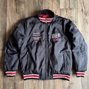 Può includere: Giacca bomber blu navy con cerniera a tutta lunghezza, finiture a righe rosse e bianche su colletto, polsini e orlo. La giacca presenta scritte e toppe ricamate, tra cui "Royal Yacht Club" e "Paul & Shark Vintage Yachting".