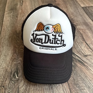 May include: Black and white trucker hat featuring the Von Dutch logo. The logo shows a winged eye and the text "Von Dutch ORIGINALS". The hat has a black peak and mesh back.