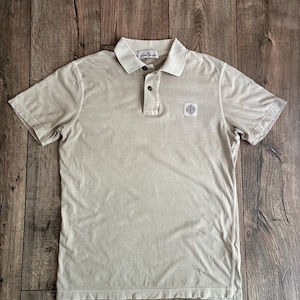Könnte beinhalten: Beiges Poloshirt mit kurzen Ärmeln, Kragen und Knopfleiste mit drei Knöpfen. Das Shirt hat einen kleinen quadratischen Aufnäher mit Logo auf der Brust. Das Shirt ist aus weichem, strukturiertem Stoff.