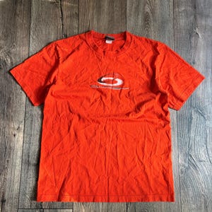 May include: Bright orange t-shirt with a crew neck and short sleeves. The shirt features a white and black graphic design on the front. The shirt is made of a soft, slightly wrinkled fabric.