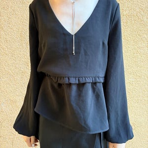 May include: Black long-sleeve top with a V-neck and flared sleeves. The top features a gathered waist detail and is paired with a matching skirt. A silver necklace with a long pendant is worn with the top.