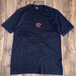 May include: Navy blue t-shirt with a small logo on the chest. The logo features text and a shark graphic in yellow, red, and white. The shirt is short-sleeved and has a crew neck.