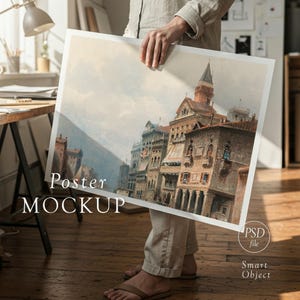 May include: A person holding a poster mockup featuring a watercolor painting of a European cityscape. The poster has a white border and the words "Poster MOCKUP" are printed on the bottom left corner. The image also includes the text "PSD file" and "Smart Object".