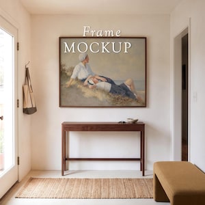 May include: A rectangular wall art frame mockup featuring a painting of two figures resting on a grassy dune by the sea. The frame is dark wood, mounted above a slim, dark wooden console table. The text Frame MOCKUP is centered above the artwork.