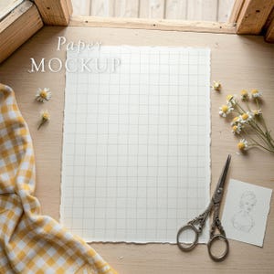May include: A mock-up of a grid paper with the text "Paper MOCKUP" on a wooden surface. The scene includes a pair of decorative scissors, a small sketch, chamomile flowers, and a yellow and white checkered cloth.