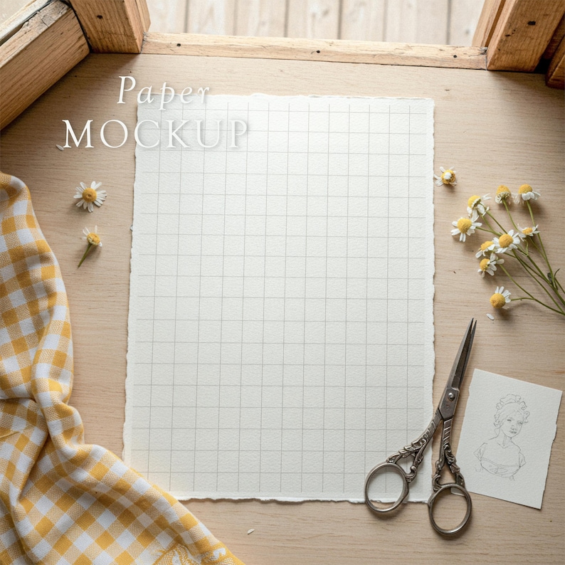 May include: A mock-up of a grid paper with the text "Paper MOCKUP" on a wooden surface. The scene includes a pair of decorative scissors, a small sketch, chamomile flowers, and a yellow and white checkered cloth.