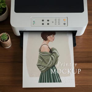 Puede incluir: Una impresora blanca imprimiendo una maqueta de una mujer con un top negro y una falda a rayas verdes, con una chaqueta verde. La impresión tiene las palabras "Printer MOCKUP" en texto blanco. La impresora está sobre una superficie de madera marrón.