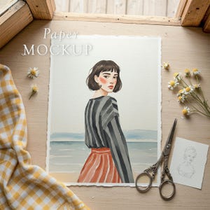 May include: Watercolor painting of a woman in a striped top and red skirt, standing near the ocean. The artwork is on textured paper, with the words "Paper MOCKUP" in the upper left corner. A pair of scissors and daisies are on the wooden surface.