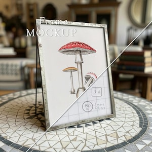 May include: A silver metal frame with a watercolor illustration of mushrooms. The frame has the text "Frame Mockup" at the top. The illustration features a large red and white mushroom, with two smaller mushrooms in the background. The frame is on a mosaic table.