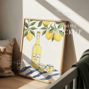 Op de afbeelding: Een ingelijste kunstdruk met een illustratie van citroenen, takken en een fles limoncello. Twee kleine mokken met citroenmotieven staan voor de fles. De lijst is van licht hout en het kunstwerk ligt op een blauw-wit gestreept oppervlak.