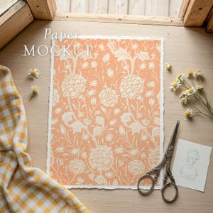 May include: A peach-colored paper with a floral pattern and the text "Paper MOCKUP" is displayed on a wooden surface. The paper is surrounded by daisies, a yellow and white checkered cloth, scissors, and a small sketch.