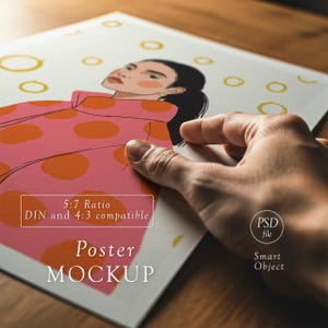以下が含まれることがあります： ピンクのコートにオレンジ色の水玉模様の女性のイラストが描かれたポスターモックアップ。ポスターには「Poster MOCKUP」と「5:7 Ratio DIN and 4:3 compatible」の文字が入っています。手でポスターを持っています。ポスターは木製の表面にあります。