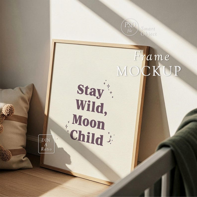 Op de afbeelding: Een ingelijste print met de tekst "Stay Wild, Moon Child" in een stijlvol lettertype. De print zit in een lichtgekleurde houten lijst en leunt tegen een muur. Een kussen en een deken zijn zichtbaar op de achtergrond.