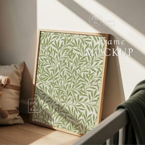 Op de afbeelding: Een ingelijste print mockup met een groen en cr&egrave;mekleurig botanisch patroon. Het houten frame leunt tegen een muur, met de tekst "Frame MOCKUP" zichtbaar. De afbeelding wordt verlicht door natuurlijk licht, wat wijst op een gezellige interieursetting.
