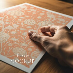 Pode incluir: Um p&ocirc;ster cor de p&ecirc;ssego com um padr&atilde;o floral, segurado por uma m&atilde;o. O design apresenta flores e folhagens brancas. As palavras "Poster MOCKUP" s&atilde;o impressas na parte inferior do p&ocirc;ster.