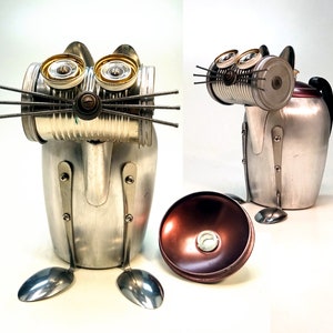 May include: A silver metal cat sculpture made from recycled materials, including a coffee pot, cans, and spoons. The cat has large, round eyes and a whimsical expression.