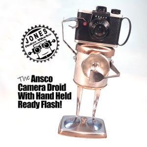 May include: A handcrafted metal robot sculpture with an Ansco camera head and a hand-held flash. The robot has a silver body, legs, and a base. The text on the image reads "The Ansco Camera Droid With Hand Held Ready Flash!"