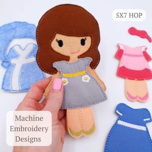 May include: A felt doll with brown hair and a gray dress with daisy accents is held in a hand. Additional felt dresses in pink and blue are visible. The text "5X7 HOP" and "Machine Embroidery Designs" are also present.