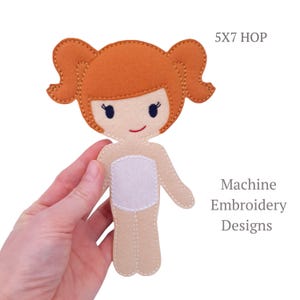 May include: A felt doll with ginger pigtails, a smiling face, and a white torso. The doll is held in a hand. The text "5X7 HOP" and "Machine Embroidery Designs" are visible.