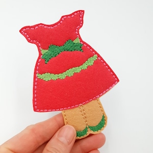 May include: Red felt dress with green felt trim and a bow. The dress has a scalloped hem and is attached to brown felt legs with green felt shoes.