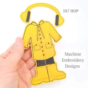 May include: Yellow felt embroidery design of a raincoat, boots, and headphones. The raincoat has black trim and buttons. The headphones have black ear pads. The text "5X7 HOP" and "Machine Embroidery Designs" are visible.