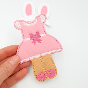 May include: A pink felt doll with a dress, bow, and shoes, with white trim. The doll has tan legs and pink shoes. White felt bunny ears are in the background. The doll is held by a hand.
