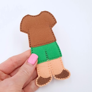 May include: A felt doll figure with a brown shirt, green shorts, tan legs, and brown shoes. The doll is hand-stitched with a contrasting thread around the edges. The figure is held against a white background.