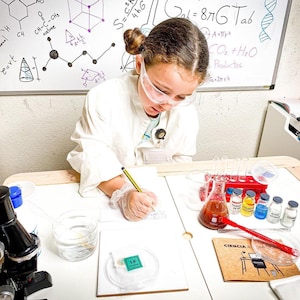 Può includere: Una giovane persona che indossa un camice da laboratorio bianco e occhiali di sicurezza sta scrivendo su un quaderno. È circondata da attrezzature scientifiche, tra cui becher, provette e un microscopio. La lavagna bianca dietro di lei ha formule e diagrammi scientifici scritti su di essa.