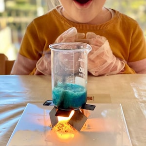 May include: A young person wearing safety goggles looks on as a beaker containing a blue liquid sits on a metal stand over a flame.
