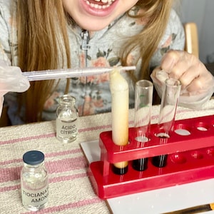 Può includere: Un bambino con occhiali e guanti di sicurezza conduce un esperimento scientifico con provette e piccoli flaconi. I flaconi sono etichettati con le parole "Ácido Cítrico" e "Tiocianato Amonio". Un supporto rosso contiene le provette.