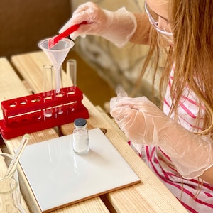 May include: A young person wearing safety goggles and gloves is conducting a science experiment. They are using a funnel to pour a white powder from a small glass bottle into a test tube. The test tube is in a red test tube rack. There is a white tile on the table.