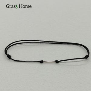 Könnte beinhalten: Schwarzes Kordelarmband mit silberfarbenem Stab. Das Armband ist schlicht und minimalistisch, mit einer dünnen, verstellbaren Kordel und einem kleinen, zylindrischen Metallakzent. Der Markenname "Grass Horse" ist sichtbar.