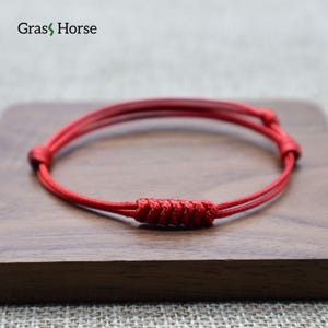 Puede incluir: Una pulsera de cuerda roja con un nudo tejido en el centro. La pulsera se muestra sobre una superficie de madera. La marca "Grass Horse" es visible en la esquina superior izquierda.