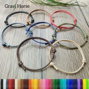 以下が含まれることがあります： 黒、茶色、青、赤など、さまざまな色の調節可能なロープブレスレット。各ブレスレットには、サイズ調整用のスライドノットが付いています。ブレスレットは木製の表面に表示され、ブランド名「Gras Horse」が見えます。