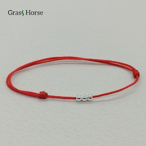 Puede incluir: Pulsera de hilo rojo con tres pequeñas cuentas plateadas. La pulsera tiene un diseño minimalista y un cierre de nudo. La marca "Grass Horse" es visible en la esquina superior izquierda.