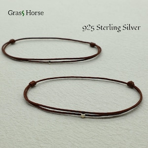 Può includere: Due braccialetti in corda marrone con una piccola perlina d'accento argentata. I braccialetti sono etichettati "925 Sterling Silver" e presentano il logo "Grass Horse". I cordoncini sono annodati a ciascuna estremità.