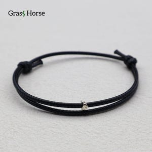 Op de afbeelding: Zwart koordarmband met een kleine zilverkleurige bedel. De armband heeft een knoopsluiting en de merknaam "Grass Horse" in de linkerbovenhoek. De armband is ontworpen met twee parallelle koorden.