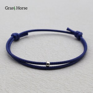 Puede incluir: Pulsera ajustable azul marino con una pequeña cuenta plateada. La pulsera está hecha de un cordón fino y tejido y tiene un cierre de nudo corredizo. La marca "Grass Horse" es visible en la esquina superior izquierda.