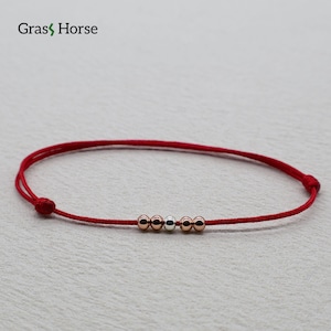 Può includere: Braccialetto con cordino rosso con quattro perline dorate e una argentata. Il braccialetto è regolabile e presenta il testo "Grass Horse". Un gioiello semplice ed elegante.