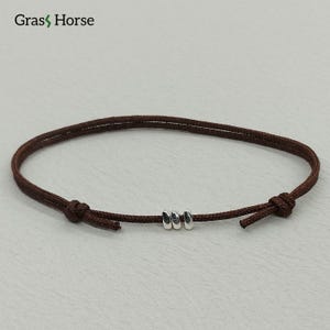 Puede incluir: Una pulsera de cordón marrón con tres pequeñas cuentas plateadas. La pulsera está anudada en cada extremo y presenta el texto "Gras Horse" en la esquina superior izquierda. Esta pulsera minimalista es adecuada para el uso diario.