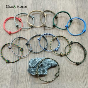 May include: A collection of colorful rope bracelets in various shades, including red, blue, green, and patterned designs. Each bracelet features a simple knot closure. The bracelets are displayed on a wooden surface.