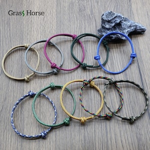 Può includere: Una collezione di braccialetti in corda colorati in varie tonalità, tra cui blu, verde e giallo. Ogni braccialetto presenta una chiusura annodata. I braccialetti sono esposti su una superficie di legno.