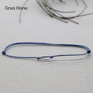 May include: A simple bracelet with a clear, spherical bead on a navy blue cord. The cord is knotted at each end and has a small silver-coloured bead. The bracelet is shown on a light grey surface.