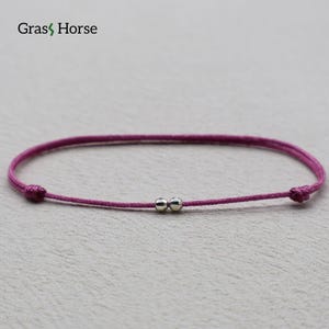 Puede incluir: Una pulsera minimalista con un cordón magenta y dos pequeñas cuentas plateadas en el centro. La pulsera está anudada en cada extremo. La marca "Grass Horse" está en la esquina superior izquierda.