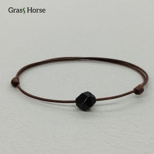 May include: A minimalist bracelet with a faceted black gemstone bead on a thin, brown cord. The cord is knotted at each end. The bracelet is designed for a simple, elegant look.